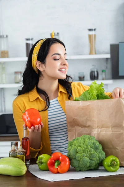 Mujer morena feliz mirando bolsa de papel con alimentos frescos mientras sostiene el pimiento - foto de stock