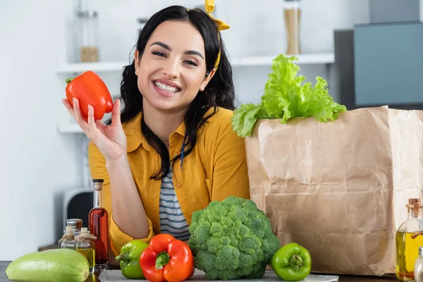 Happy brunette woman smiling near paper bag with fresh groceries while holding bell pepper — Stock Photo