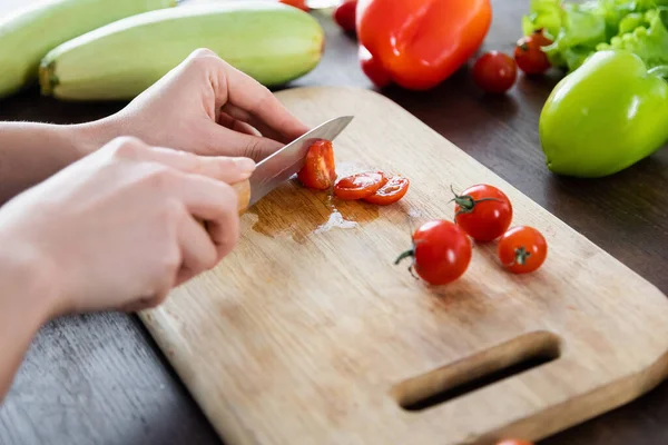 Vista recortada de la mujer cortando tomates cherry en la tabla de cortar cerca de verduras - foto de stock