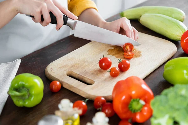 Vista ritagliata della donna che taglia pomodorini ciliegini sul tagliere di legno vicino alle verdure — Foto stock