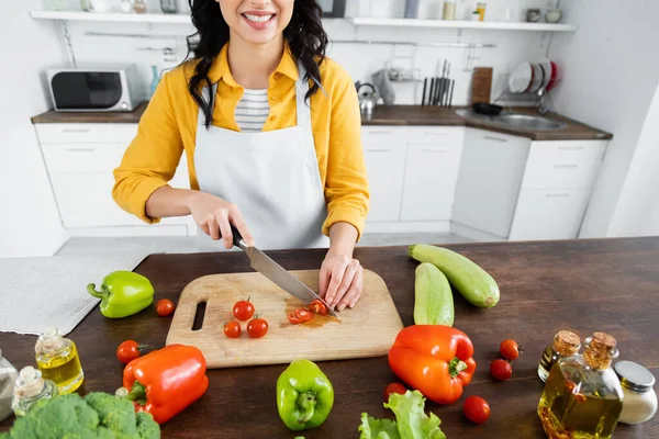 Vista cortada de mulher alegre cortando tomates cereja em tábua de corte de madeira perto de legumes — Fotografia de Stock