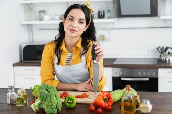 Young brunette woman holding knife near wooden chopping board and vegetables on kitchen table — Stock Photo