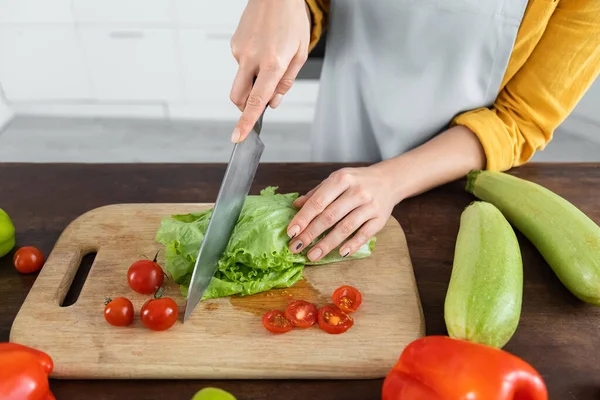Cropped view of woman cutting fresh lettuce near red cherry tomatoes on wooden chopping board — Stock Photo