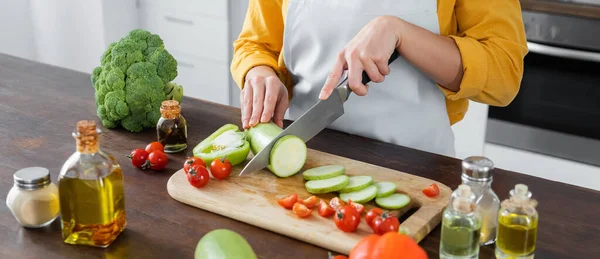 Vista recortada de la mujer en delantal cortando calabacín cerca de tomates cherry en la tabla de cortar, pancarta - foto de stock