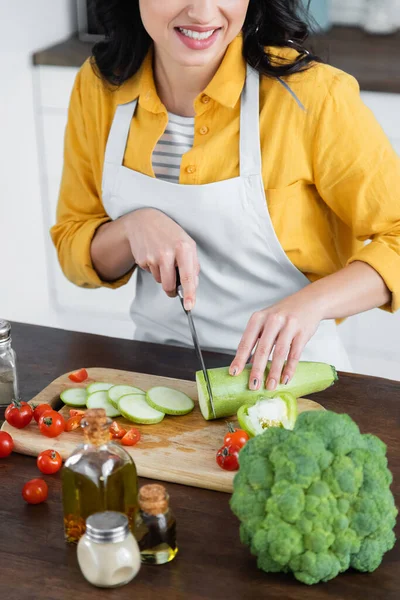 Обрезанный вид улыбающейся женщины в фартуке, режущей цуккини рядом с помидорами черри на доске — стоковое фото