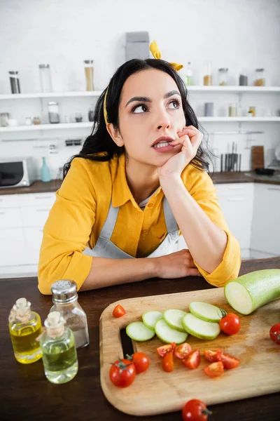 Mujer joven pensativa mirando hacia otro lado mientras piensa cerca de verduras en primer plano borrosa - foto de stock