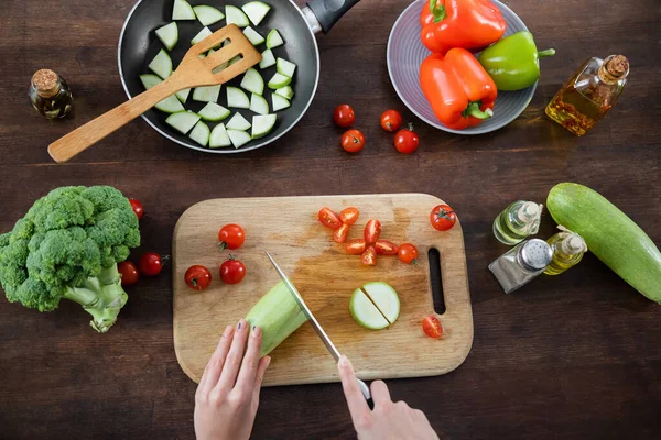 Draufsicht einer Frau, die Zucchini in der Nähe von in Scheiben geschnittenen Kirschtomaten auf Schneidebrett und Pfanne auf dem Tisch schneidet — Stockfoto