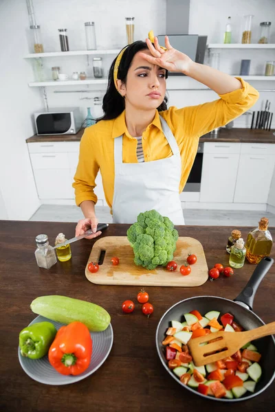 Tired young woman in apron wiping sweat while holding knife near vegetables and frying pan in kitchen — Stock Photo