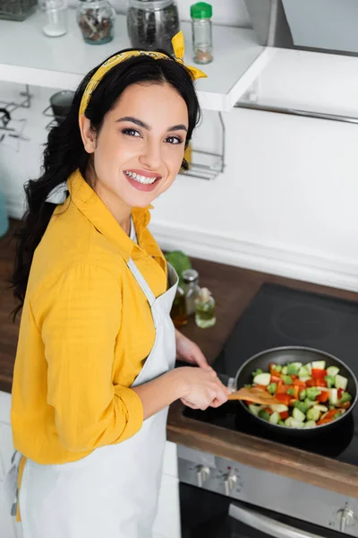 Vista de ángulo alto de la mujer morena feliz mezclando verduras en rodajas en la sartén con espátula - foto de stock