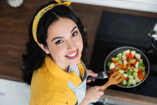 Vista de ángulo alto de la mujer morena alegre mezcla de verduras en rodajas en la sartén con espátula - foto de stock