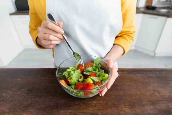 Vista recortada de mujer joven en delantal sosteniendo tenedor cerca de ensalada en tazón - foto de stock