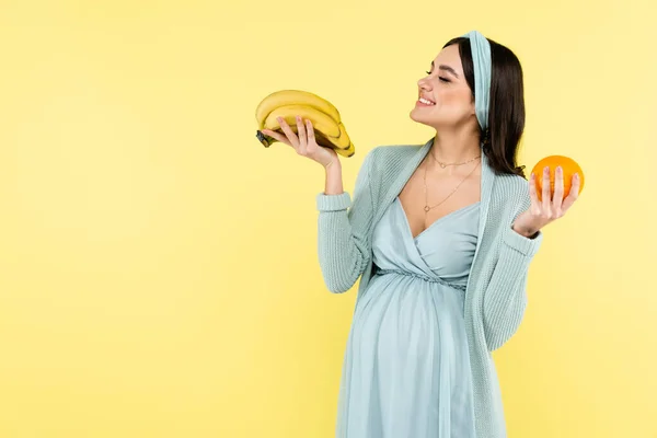 Mujer embarazada alegre sosteniendo plátanos maduros aislados en amarillo - foto de stock