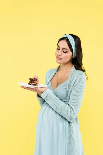 Junge schwangere Frau zweifelt, während sie leckeren Kuchen auf gelb isoliert hält — Stockfoto