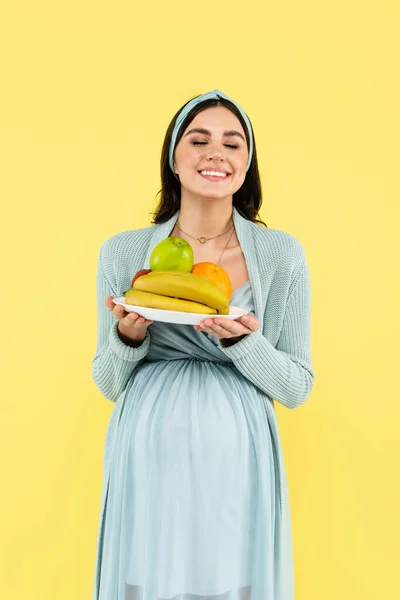 Happy pregnant woman with closed eyes holding plate with fresh fruits isolated on yellow — Stock Photo