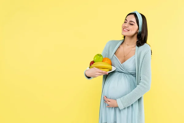 Mulher grávida sorrindo segurando prato com frutas maduras isoladas no amarelo — Fotografia de Stock