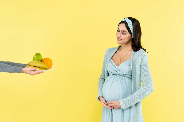 Lächelnde Schwangere, die den Bauch berührt, während sie frische Früchte sieht, die auf gelb isoliert sind — Stockfoto