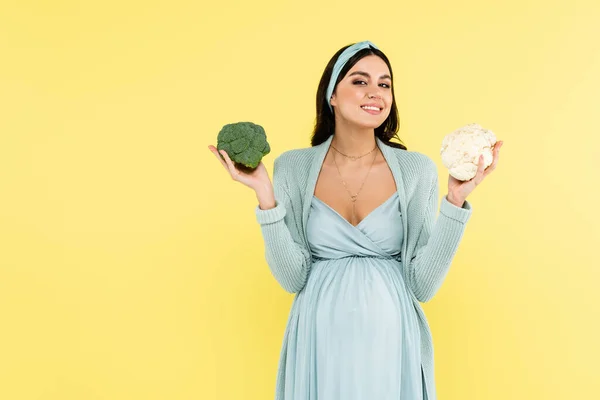 Junge schwangere Frau mit Brokkoli und Blumenkohl lächelt isoliert auf gelb in die Kamera — Stockfoto