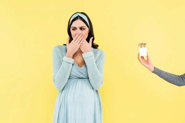 Pregnant woman feeling disgust and covering mouth near cigarette pack isolated on yellow — Stock Photo