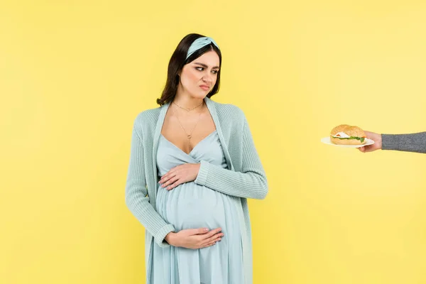 Pregnant woman feeling disgust while looking at burger isolated on yellow — Stock Photo