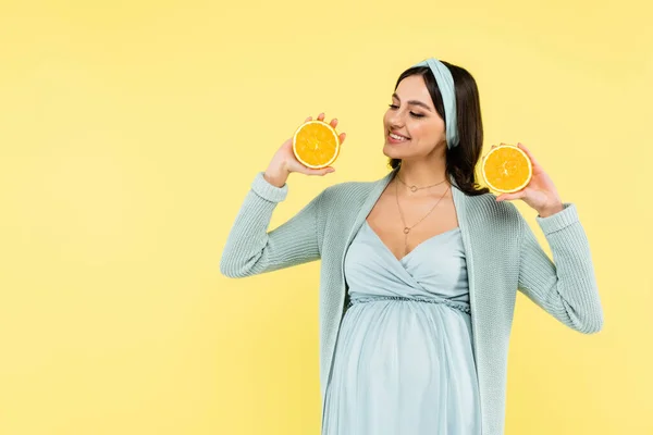 Mujer embarazada alegre sosteniendo mitades de naranja madura aisladas en amarillo - foto de stock