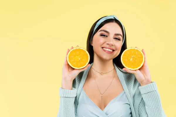 Jeune femme souriant à la caméra tout en tenant des moitiés d'orange fraîche isolé sur jaune — Photo de stock