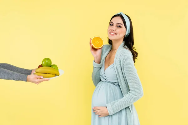 Pleased pregnant woman holding half of juicy orange near ripe fruits isolated on yellow — Stock Photo