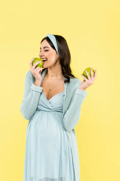 Happy pregnant woman with closed eyes eating juicy apple isolated on yellow — Stock Photo