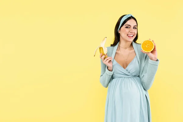 Feliz embarazada sonriendo a la cámara mientras sostiene plátano y jugoso naranja aislado en amarillo - foto de stock