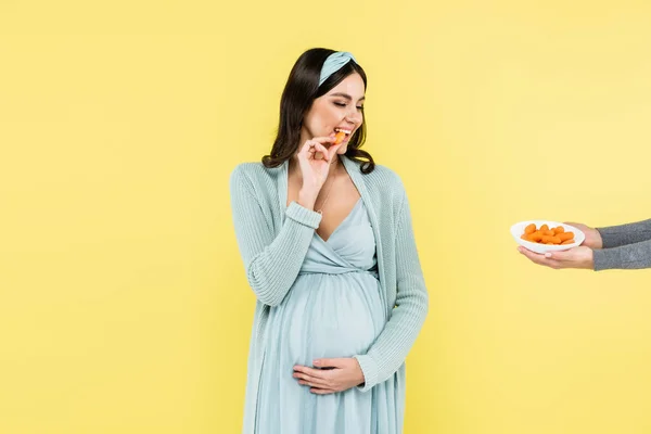 Fröhliche schwangere Frau blickt auf Schüssel mit frischer Karotte auf gelb isoliert — Stockfoto