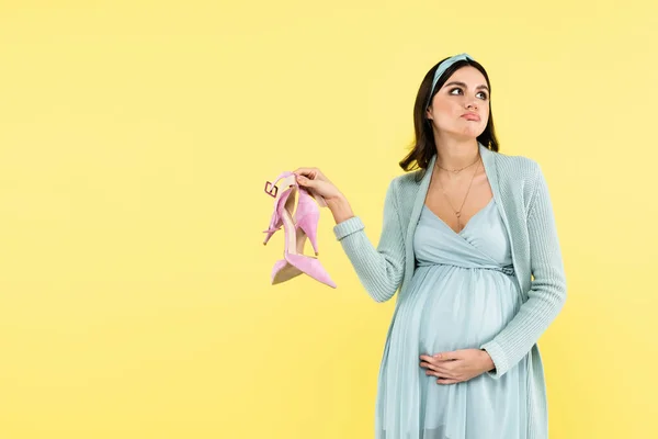 Mujer embarazada reflexiva sosteniendo zapatos de tacón alto aislados en amarillo - foto de stock