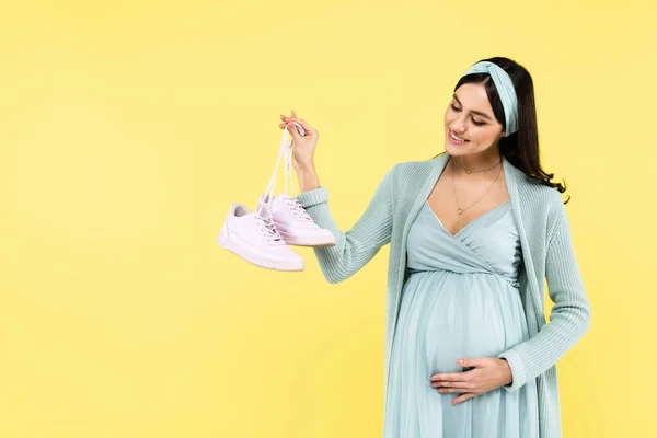 Joyful pregnant woman touching belly while holding sneakers isolated on yellow — Stock Photo