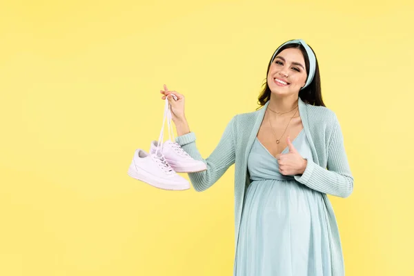 Happy pregnant woman showing thumb up while holding sneakers isolated on yellow — Stock Photo