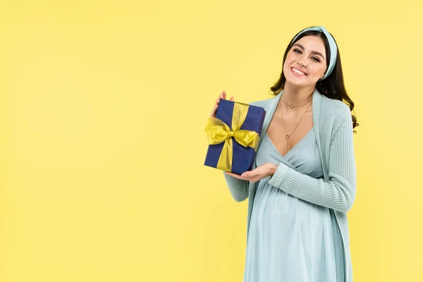 Веселая беременная женщина улыбается в камеру, держа в руках подарок, изолированный на желтый — стоковое фото