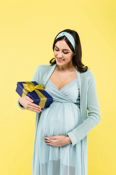Pleased pregnant woman touching tummy while holding present isolated on yellow — Stock Photo