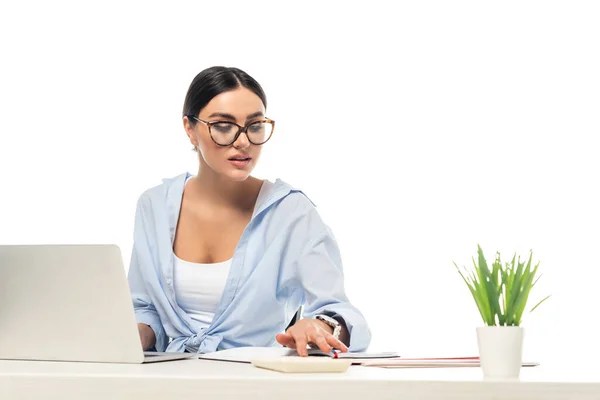 Jeune femme d'affaires en utilisant une calculatrice tout en travaillant près d'un ordinateur portable isolé sur blanc — Photo de stock