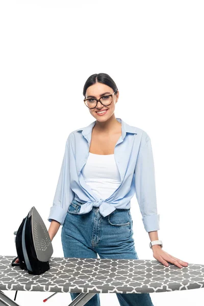 Young woman in eyeglasses smiling at camera near iron and ironing board isolated on white — Stock Photo