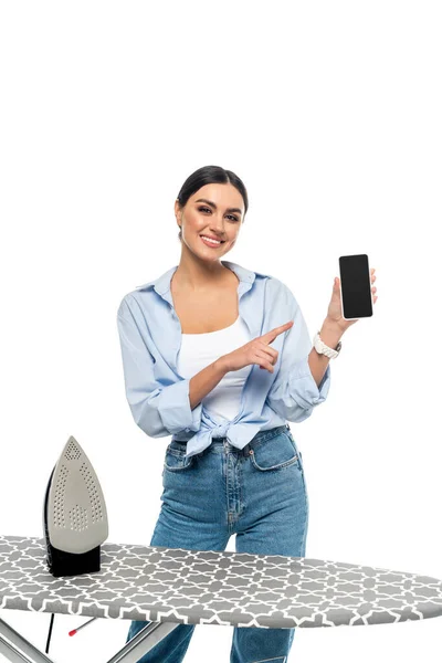 Ama de casa feliz apuntando al teléfono inteligente con pantalla en blanco cerca de tabla de planchar aislado en blanco - foto de stock