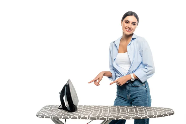 Smiling woman pointing at iron on ironing board isolated on white — Stock Photo