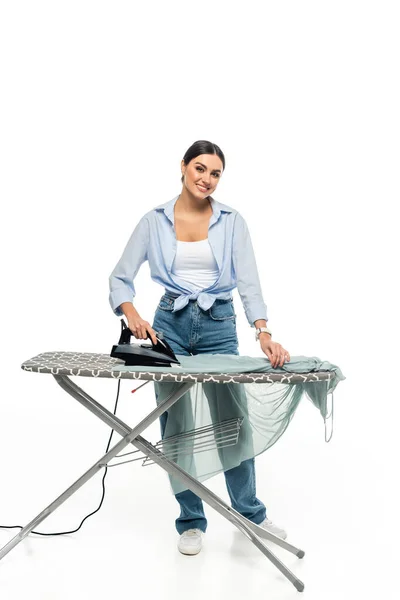 Mujer alegre sonriendo a la cámara mientras plancha la ropa sobre fondo blanco — Stock Photo