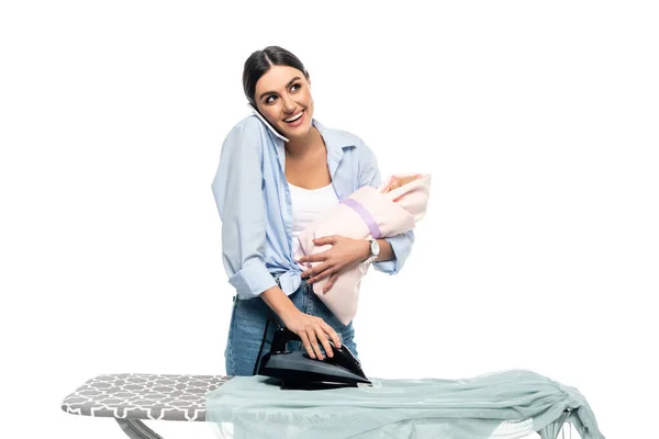 Mulher feliz segurando bebê e falando no telefone celular enquanto engomando roupas isoladas no branco — Fotografia de Stock