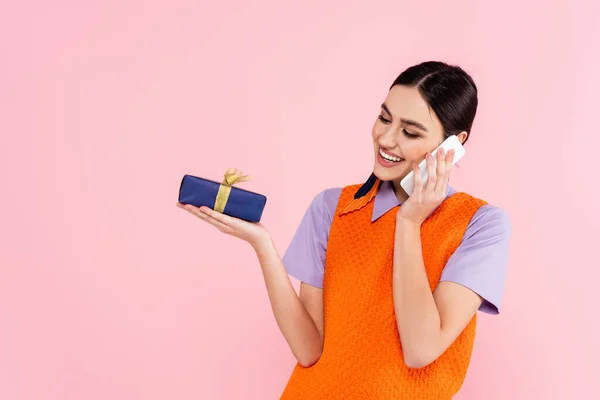 Cheerful woman talking on cellphone while holding present isolated on pink — Stock Photo