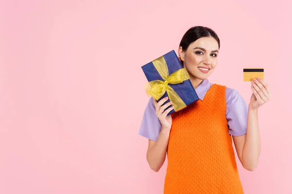 Pretty woman smiling at camera while holding credit card and present isolated on pink — Stock Photo