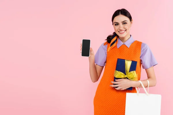 Felice donna che mostra il telefono cellulare con schermo bianco mentre tiene i regali isolati su rosa — Foto stock