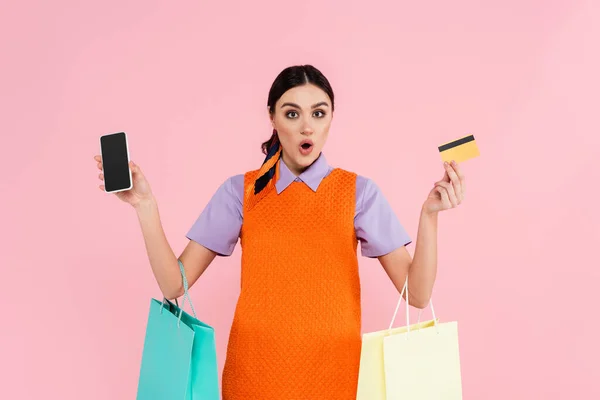 Erstaunte Frau zeigt Smartphone und Kreditkarte, während sie Einkaufstüten isoliert in rosa hält — Stockfoto