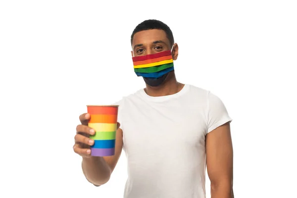 Homme afro-américain avec masque de sécurité et tasse en papier aux couleurs arc-en-ciel isolé sur blanc, concept lgbt — Photo de stock