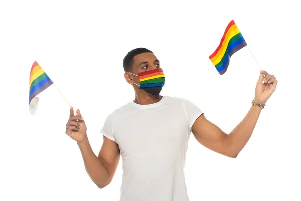 Uomo afro-americano che tiene bandiere lgbt mentre indossa colori arcobaleno maschera medica isolata su bianco — Foto stock