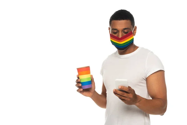 Hombre afroamericano con máscara protectora y taza de papel en colores lgbt mensajería en el teléfono inteligente aislado en blanco - foto de stock