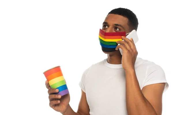Hombre afroamericano con máscara médica y taza de papel en colores lgbt hablando en el teléfono celular aislado en blanco - foto de stock