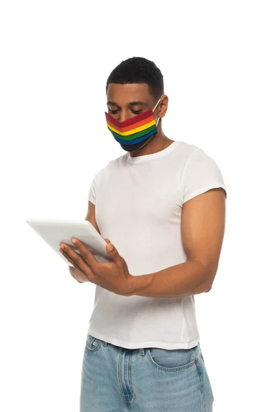 Hombre afroamericano con máscara de seguridad en colores lgbt y el uso de tableta digital aislado en blanco - foto de stock