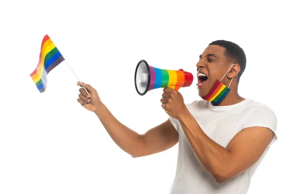 Uomo afroamericano con i colori dell'arcobaleno maschera medica tenendo bandiera lgbt e urlando in altoparlante isolato su bianco — Foto stock